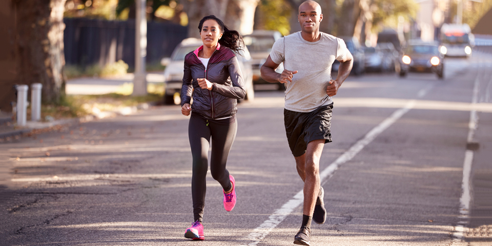 
A man and a woman jog side by side on a city street, both dressed in athletic wear. Their focused expressions and steady pace highlight their commitment to adopting an active lifestyle through regular running. The urban environment with trees and traffic in the background suggests they are making fitness a priority in their everyday routine.