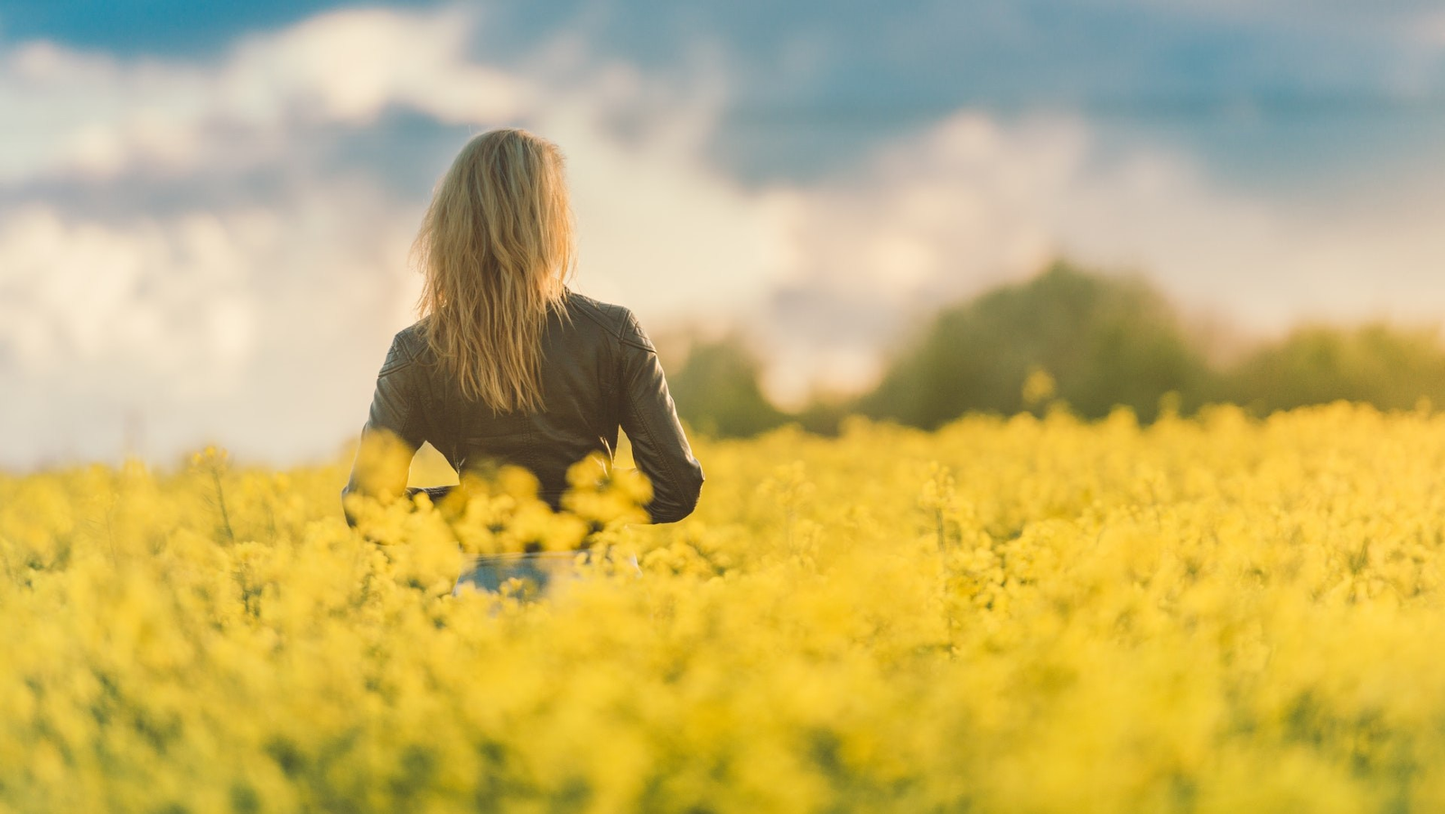 Woman in leather jacket standing in a field of yellow flowers with a sunset sky.