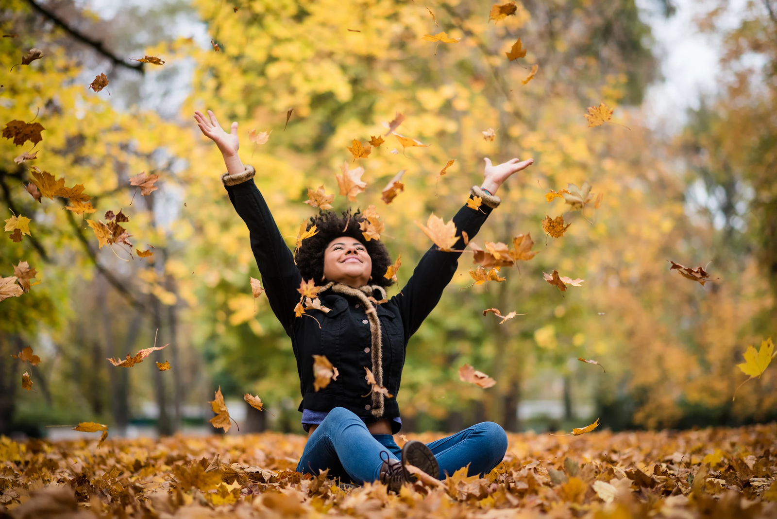 Person sitting on a carpet of autumn leaves with arms raised, surrounded by falling foliage.