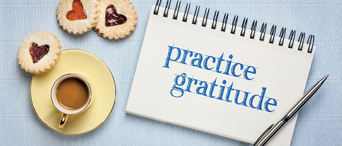 Notebook with "practice gratitude" text, espresso cup, pen, and heart-shaped cookies on blue background.