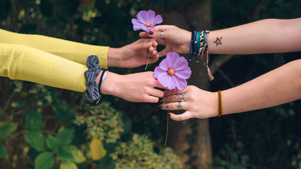 Four hands exchanging purple flowers against a natural green backdrop.