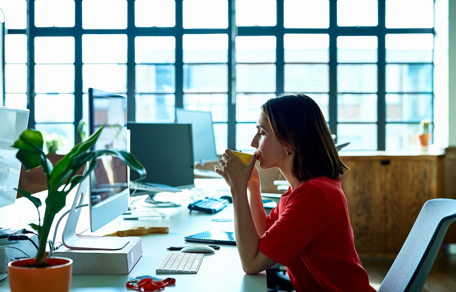 Person in red shirt at a desk with computers holding a cup in an office with large windows.