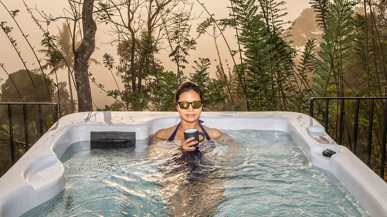 Person in a hot tub holding a mug with a forested background.