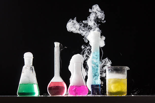 Five lab flasks with colored liquids emitting smoke on a dark background.