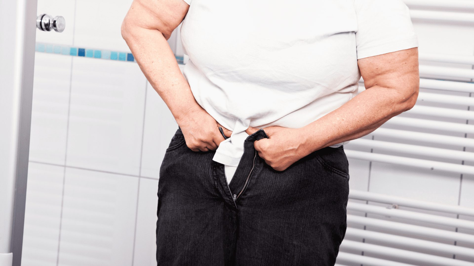 Person struggling to fasten jeans against a white bathroom background.