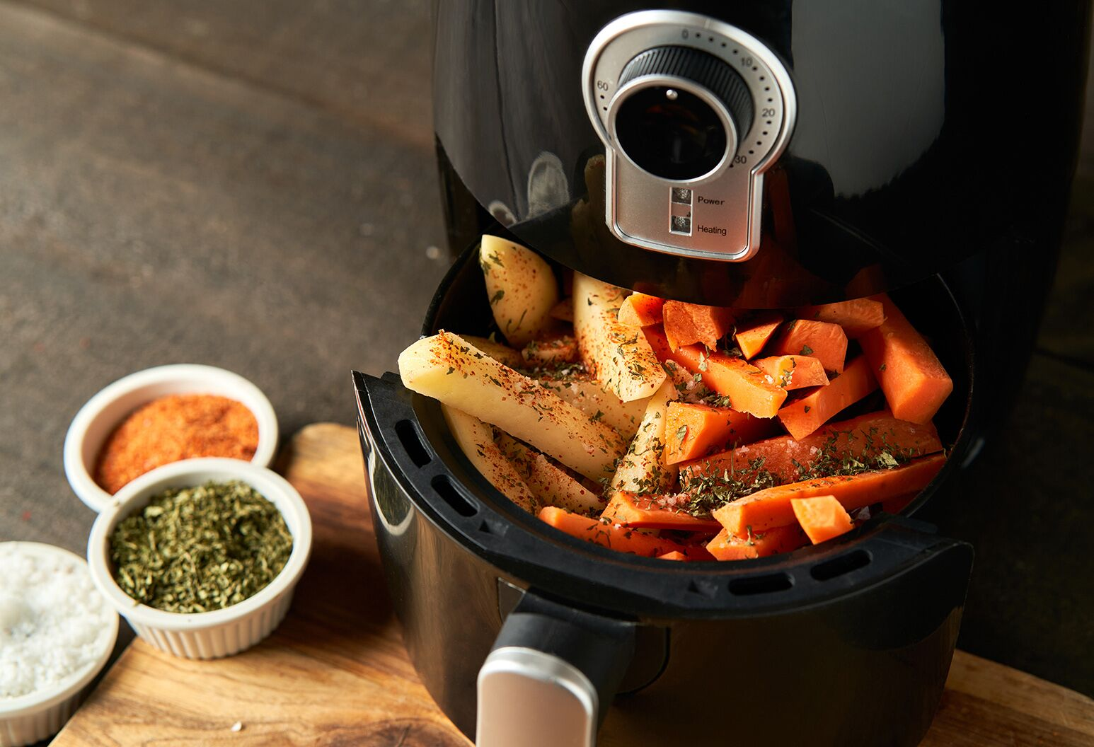 Air fryer with seasoned potato and sweet potato wedges, and spice bowls nearby.