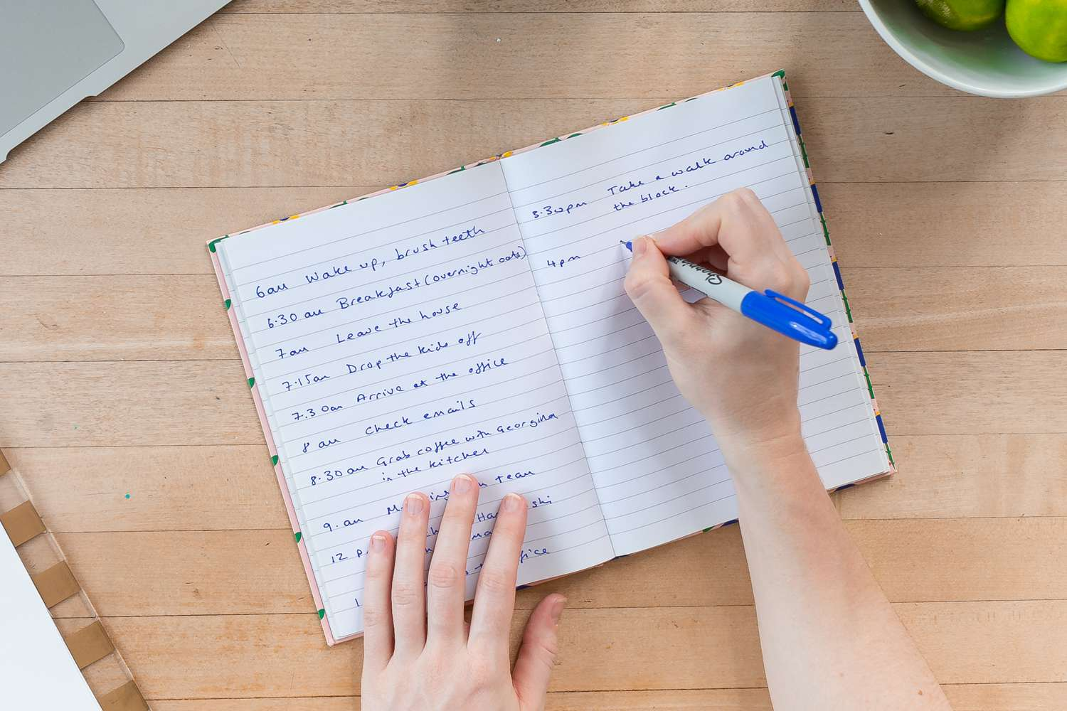 Hand writing a schedule in a notebook with a laptop and green apples in the background.