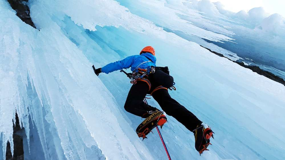 Climber ascending a steep icy slope with ice axes and crampons.