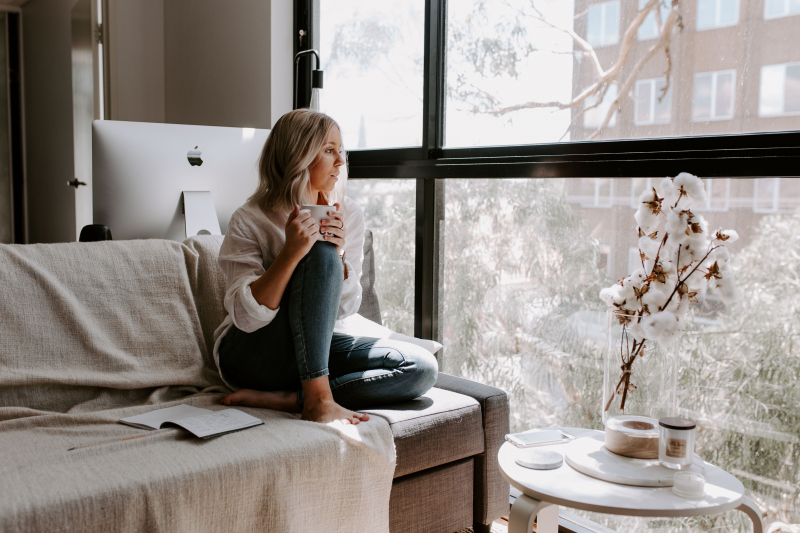 Person sitting by a window holding a mug, with an open book and a vase with branches on the table.