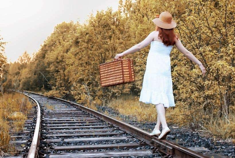 Woman in white dress and hat with basket walking on train tracks amidst trees.