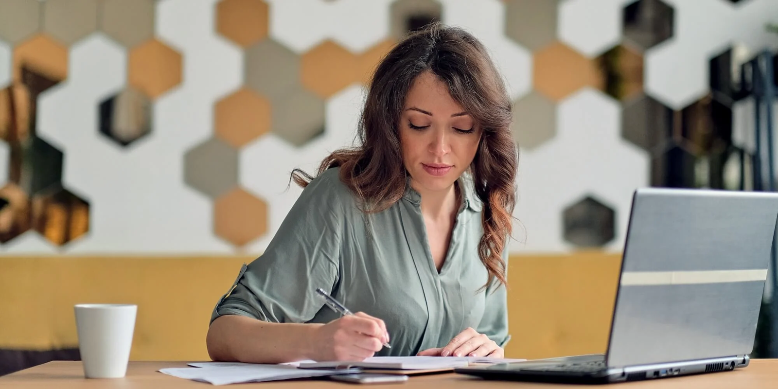 Woman writing on paper beside a laptop on a desk with a coffee cup.